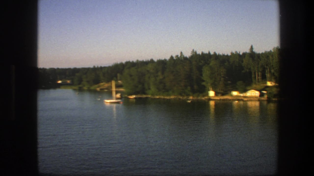 1975 Swedish 8mm view of a calm lake, small sailboat, wooded shoreline, and lakeside cabins seen through dark window framing.