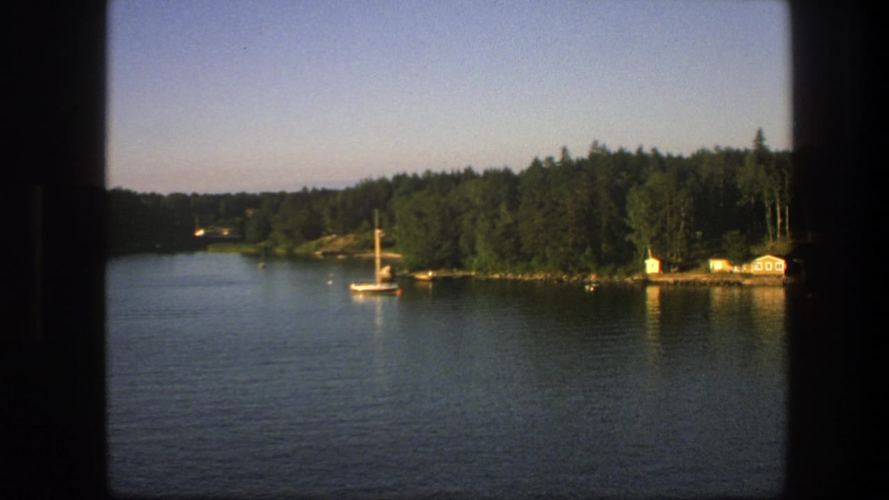 1975 Swedish lakeside home-movie frame with a sailboat, dense trees, small shoreline buildings, and warm reflections on the water.