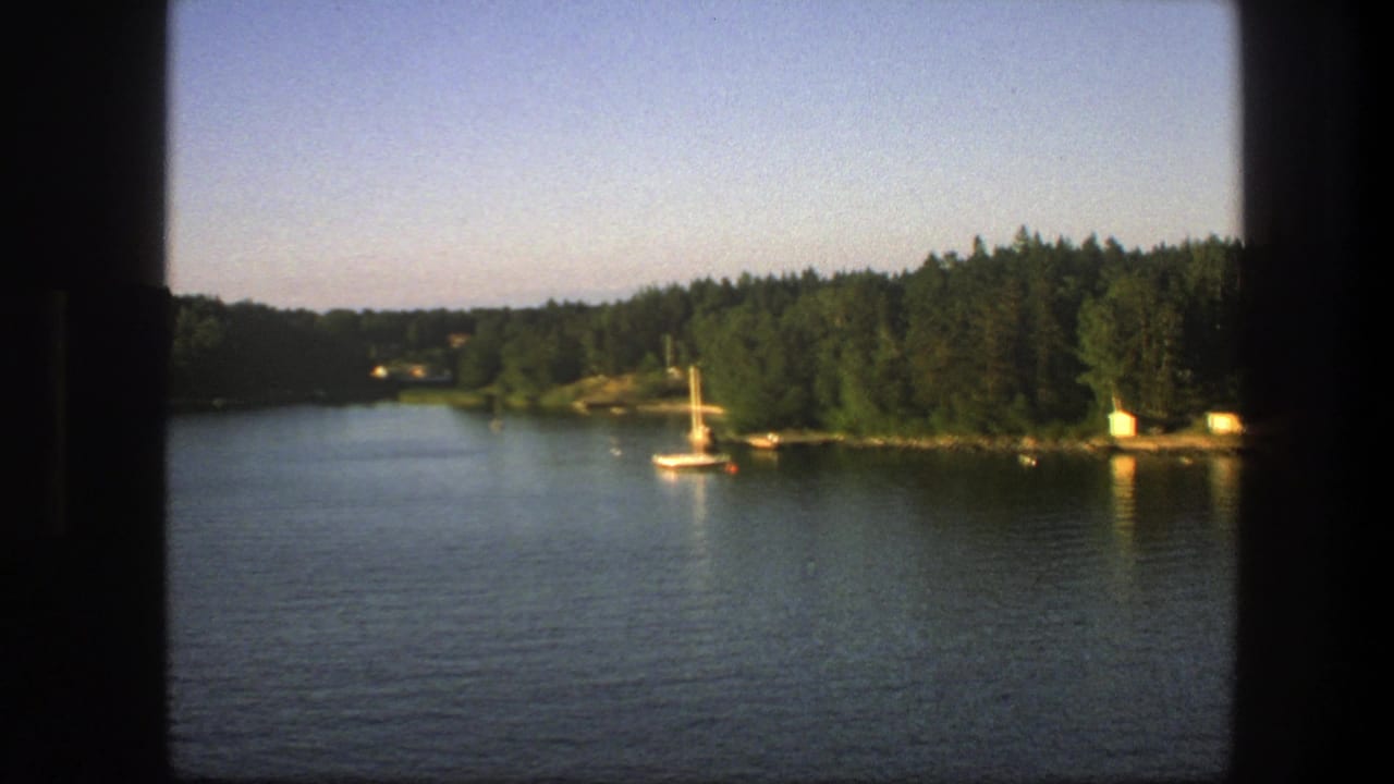 1975 Swedish 8mm frame showing a wide calm lake, sailboat near shore, dense conifer trees, and dark window framing in the foreground.