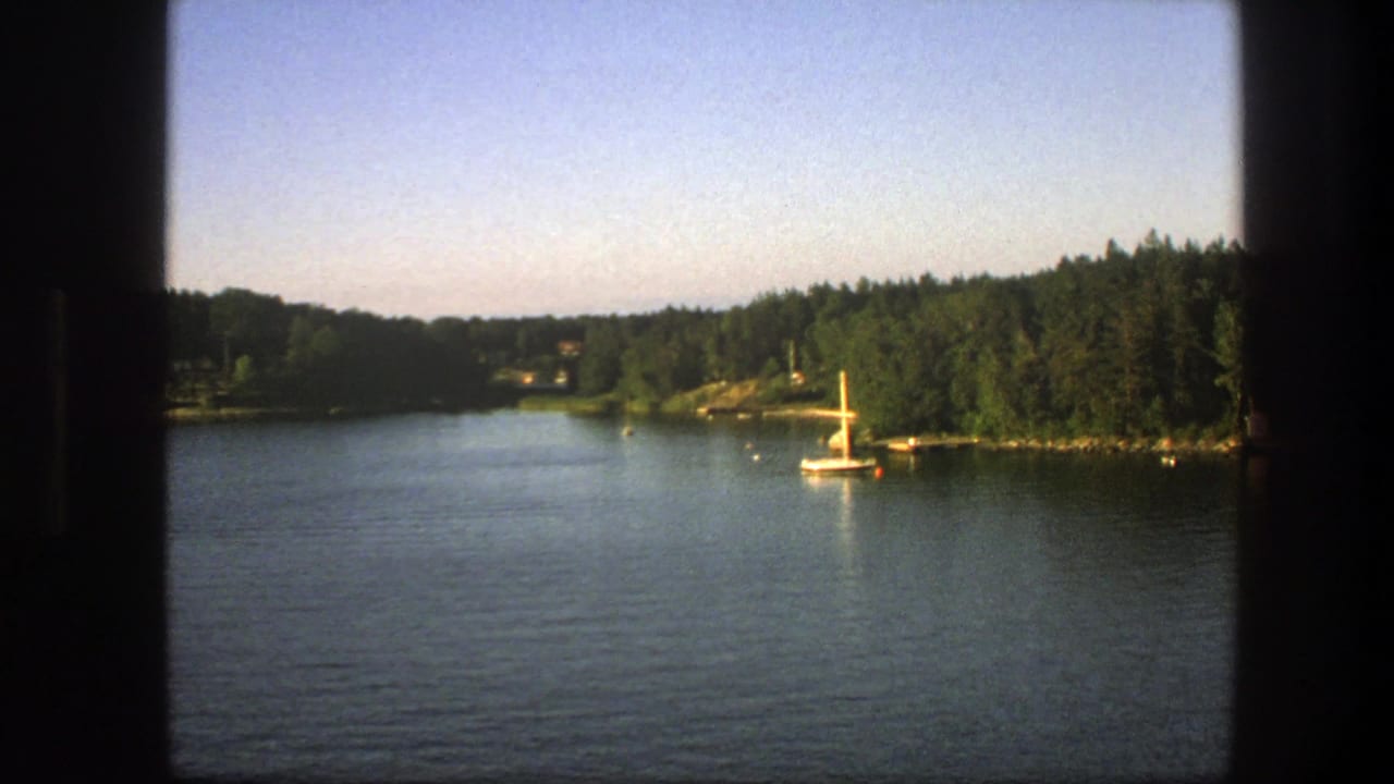 1975 Swedish home-movie still of a sailboat on calm water near a wooded shoreline, with small structures and soft dusk light.
