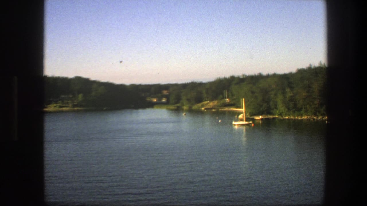 1975 Sweden 8mm view of a white sailboat on calm blue water near a tree-lined shore, framed by a dark window edge.