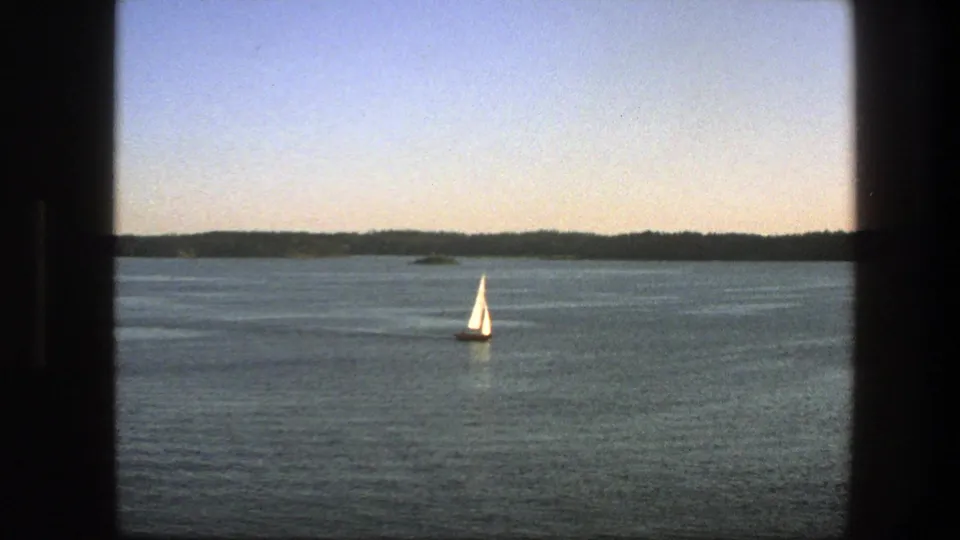 1975 Sweden 8mm frame showing a white sailboat crossing open blue water below a distant tree-lined horizon.