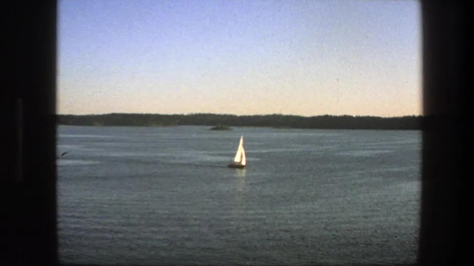 1975 Sweden 8mm view of a white sailboat on calm water, with low wooded shoreline and pale sky behind it.