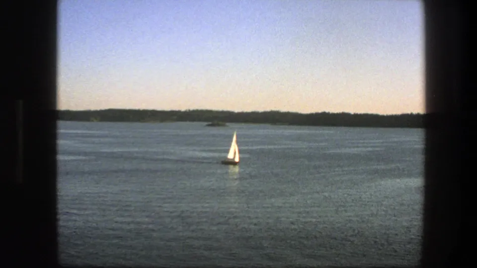 1975 Sweden 8mm frame of a white sailboat on calm blue water, bordered by dark window framing and a wooded horizon.