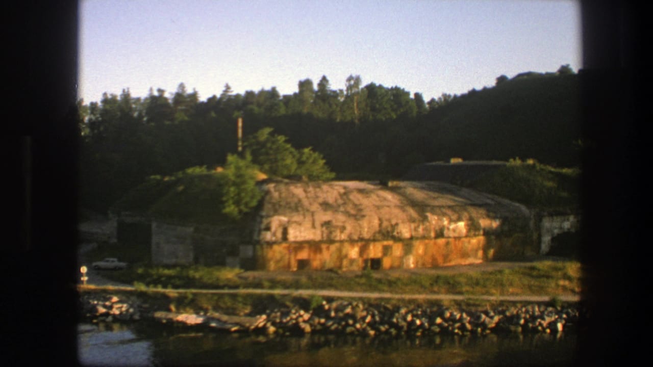 1975 Sweden 8mm frame showing a weathered concrete bunker near a rocky shoreline, with forest and green hills in the background.