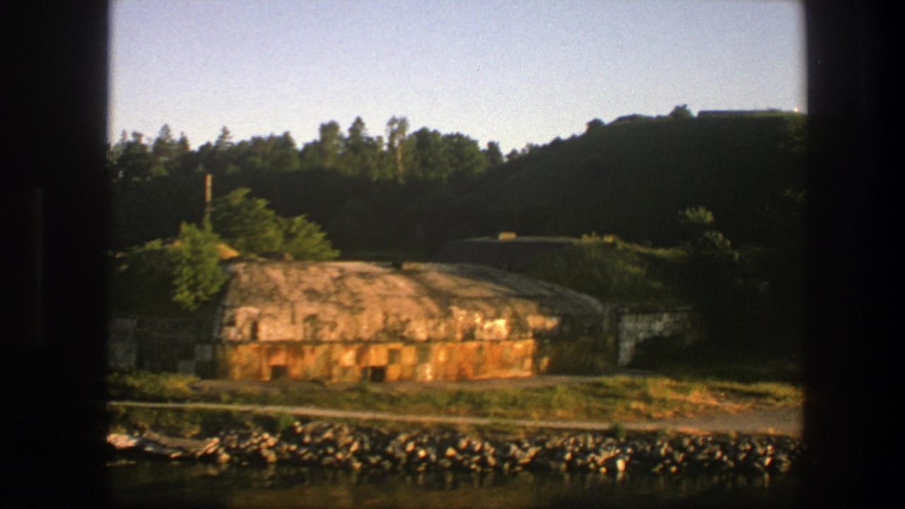 Weathered concrete bunker near a rocky water edge with hills and trees behind.