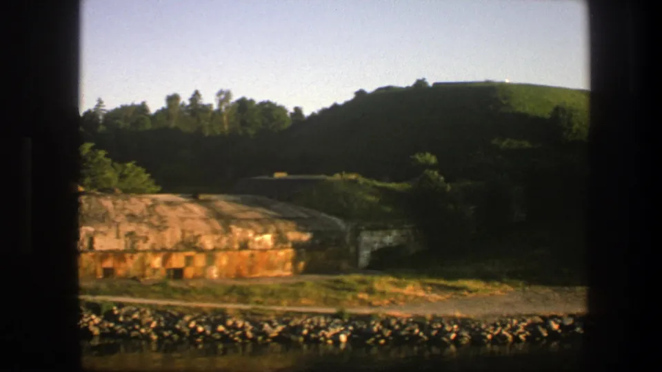 1975 Sweden 8mm frame with the bunker partly cropped as the camera moves past a rocky shoreline and forested hillside.