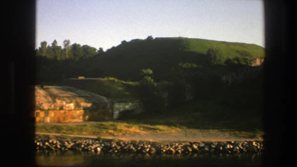 Late shot from the 1975 Sweden 8mm clip showing the bunker receding near water, with rocks, trees, and a green hillside behind.