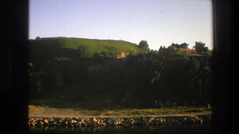 1975 Sweden 8mm view across a rocky river edge toward a green hillside, dense trees, low brush, and a small structure-like stone feature.