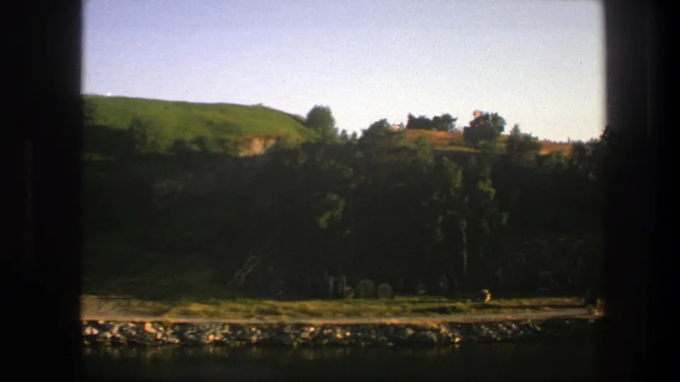 1975 Sweden frame showing a green hillside, clustered trees, rocky riverside ground, pale sky, and dark vertical window framing in warm 8mm color.