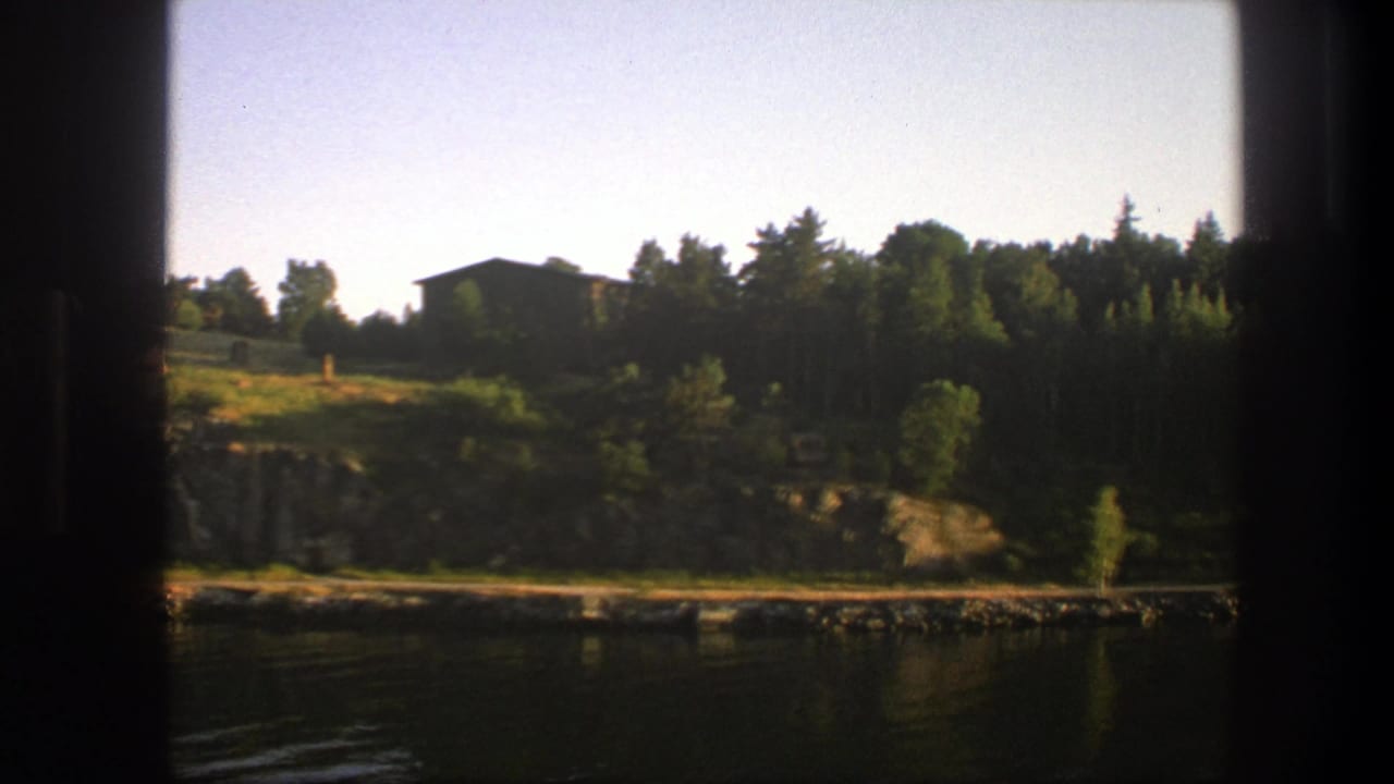 1975 8mm view across still water toward a wooded Swedish shoreline with rocks, grass, trees, and a small dark cabin.