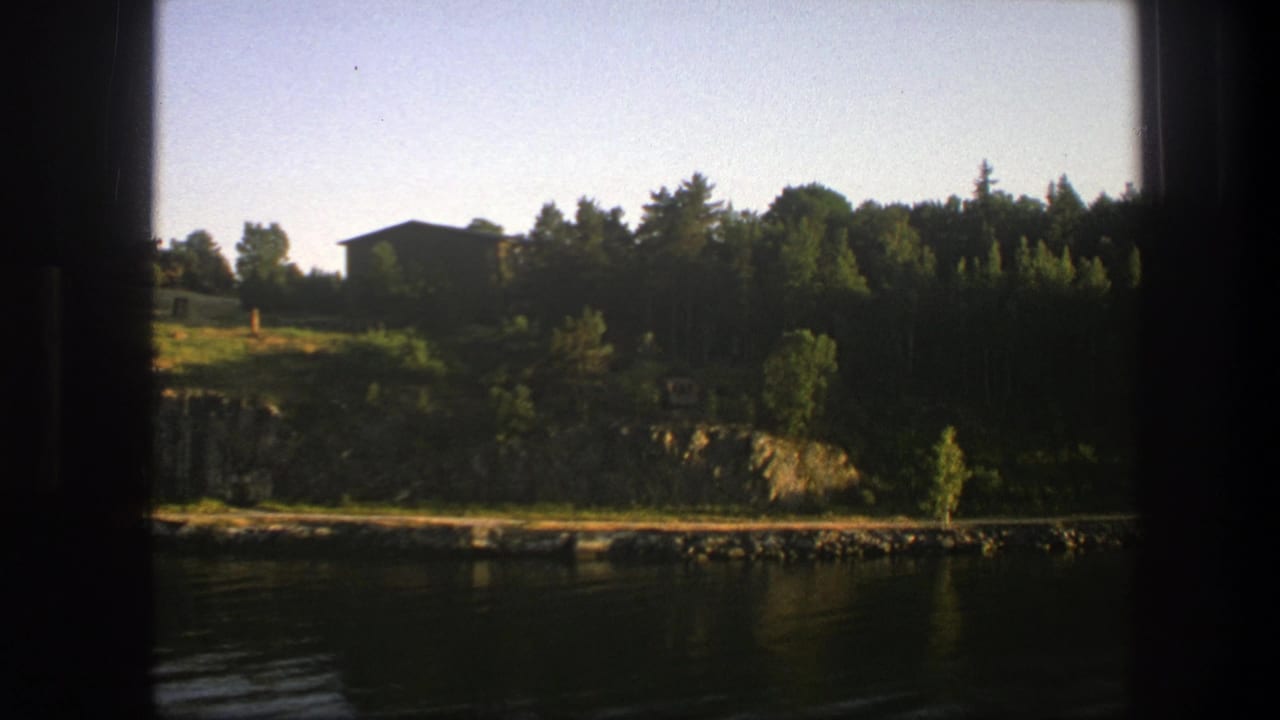 Still water below a rocky, tree-lined shore with a dark cabin set back on the hill.
