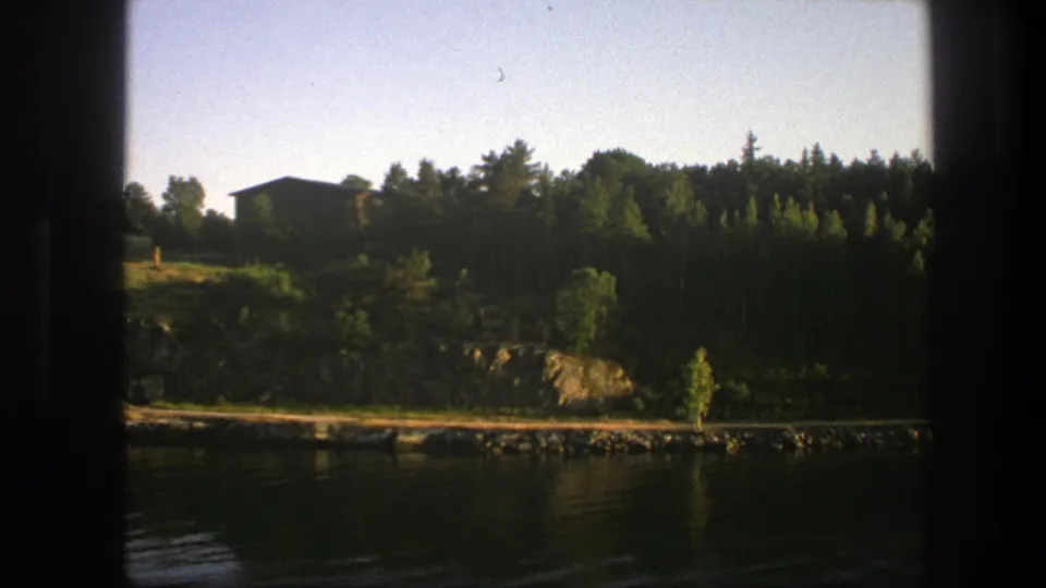 1975 Swedish home-movie frame of a quiet wooded shoreline, dark water, sunlit rocks, and a small cabin near the treeline.