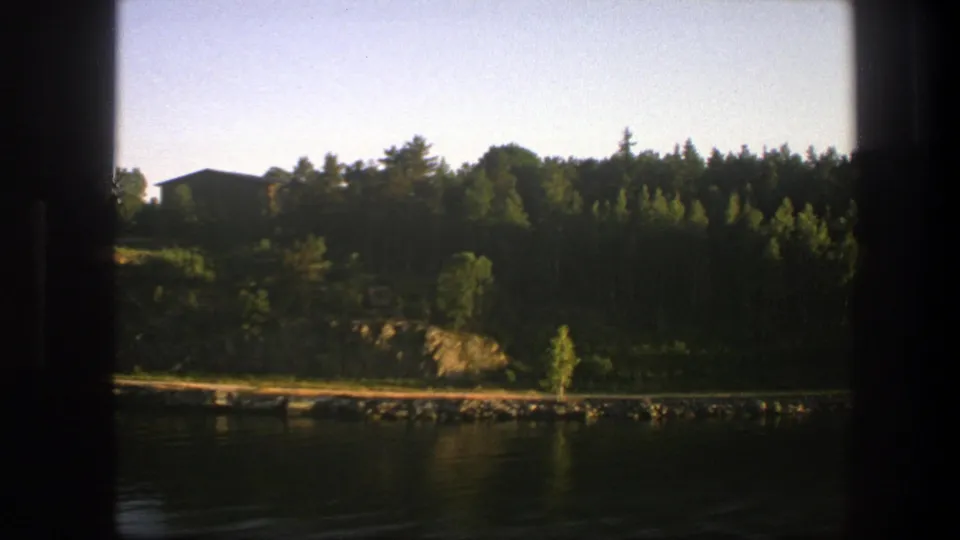 Late-frame 8mm view across calm Swedish water to a rocky, forested shore with a small dark cabin visible above the bank.