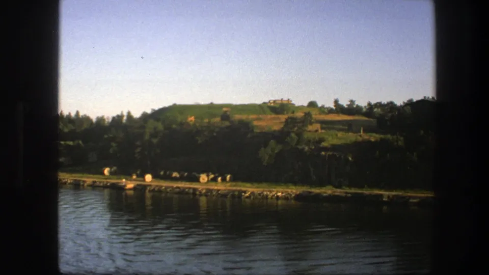 Swedish lakeside view with rippled water, wooded bank, open hillside, and small distant structures on the ridge.