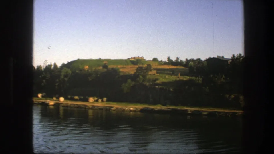1975 Swedish home-movie frame showing water, a wooded bank, hillside fields, and distant buildings through dark 8mm edges.