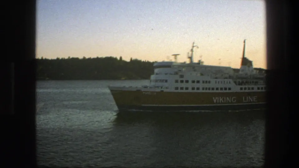 A yellow and white Viking Line ferry passes across calm water near a wooded Swedish shoreline in 1975 home-movie footage.