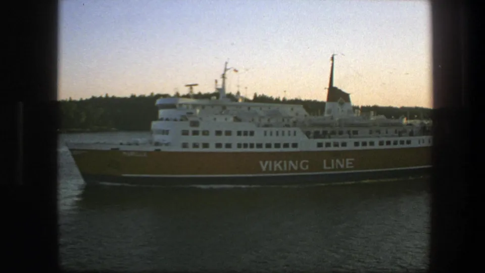 The branded passenger ferry continues past the camera on calm water, framed by wooded shoreline and soft 1970s 8mm color.