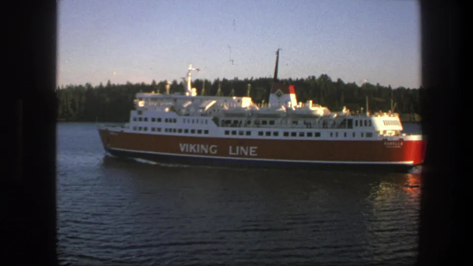 1975 Sweden 8mm frame of a red-and-white Viking Line ferry moving across calm water near a tree-lined shore.