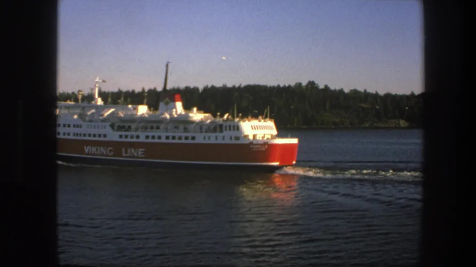 1975 Sweden 8mm view of the ferry's stern and branded hull as it passes, leaving a pale wake behind.