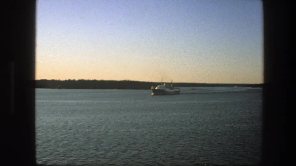 1975 Sweden 8mm frame showing a ferry moving across calm lake water below a pink-blue evening sky.