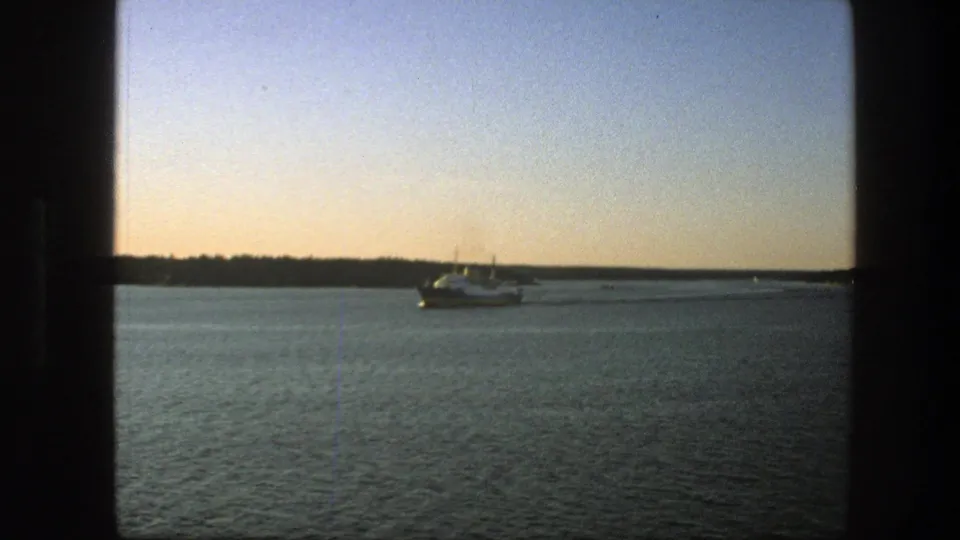 1975 home-movie frame of a ferry on a Swedish lake, with dusk color and dark interior framing at the edges.