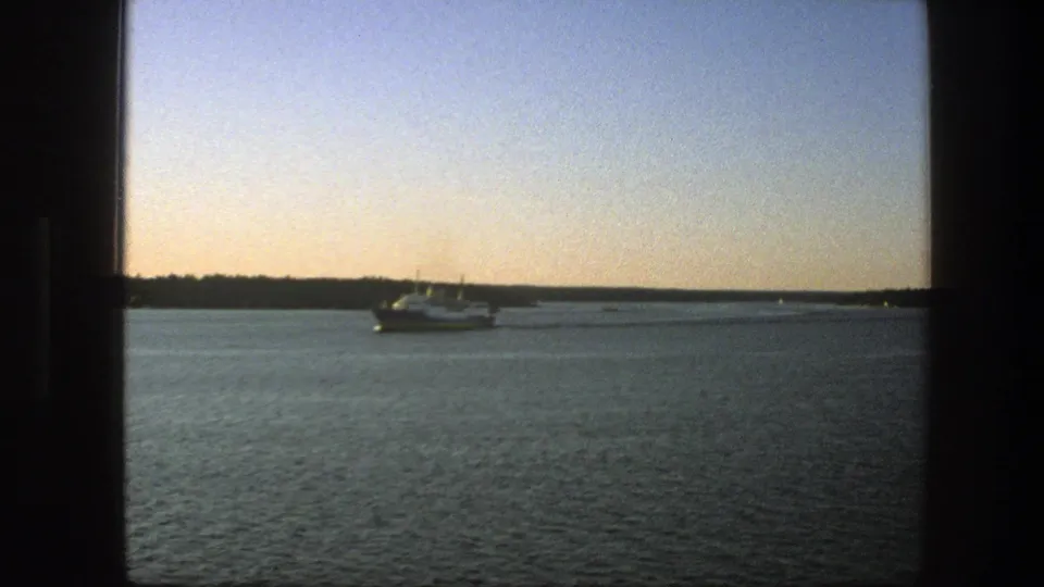 Swedish 1975 8mm still of a ferry crossing calm water at dusk, with low shoreline and muted sky colors.