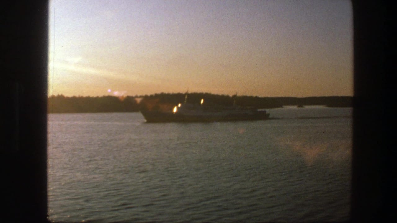 Large ship in silhouette on calm water at sunset, with small lights visible near the hull.