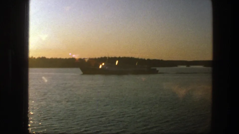 1975 Sweden 8mm frame of a large ship moving along calm water, partly silhouetted against a hazy golden shoreline.