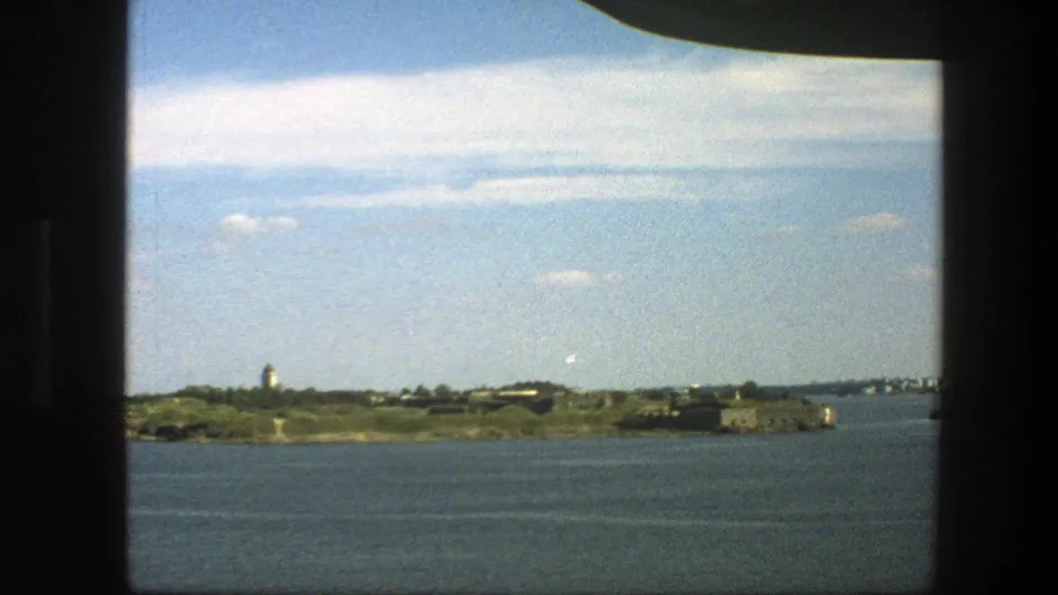1975 Sweden 8mm frame showing calm water, a distant island or shoreline, and a small tower-like structure beneath a partly cloudy sky.