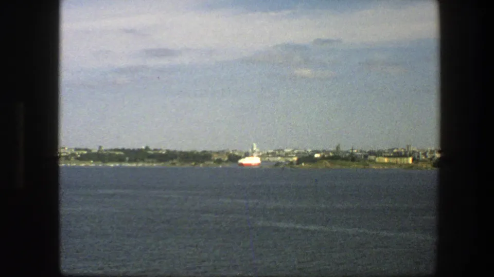 1975 Sweden 8mm frame showing a distant waterfront, industrial-looking low structures, and a red-and-white ship on the horizon.