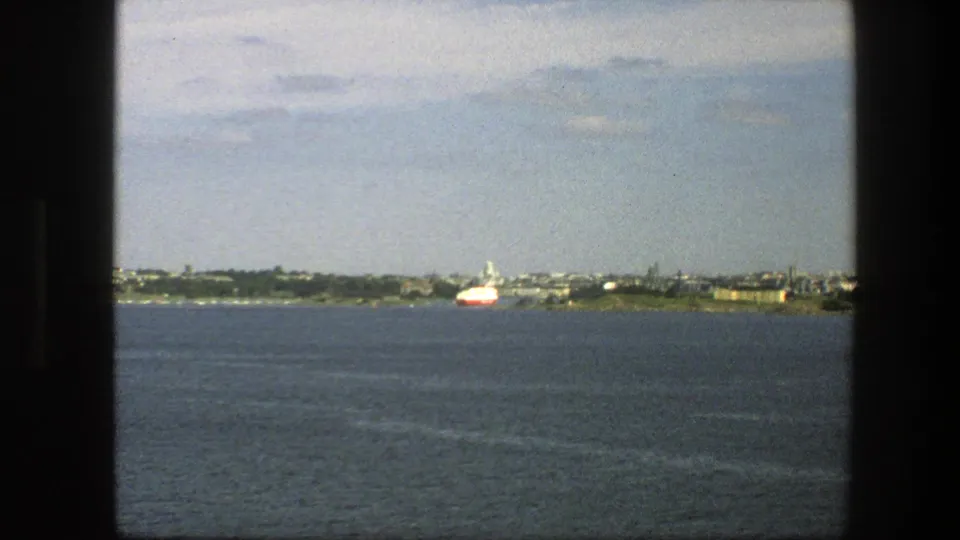 1975 Sweden 8mm view from a moving vessel, with open water, distant coastal buildings, and a red and white ship.