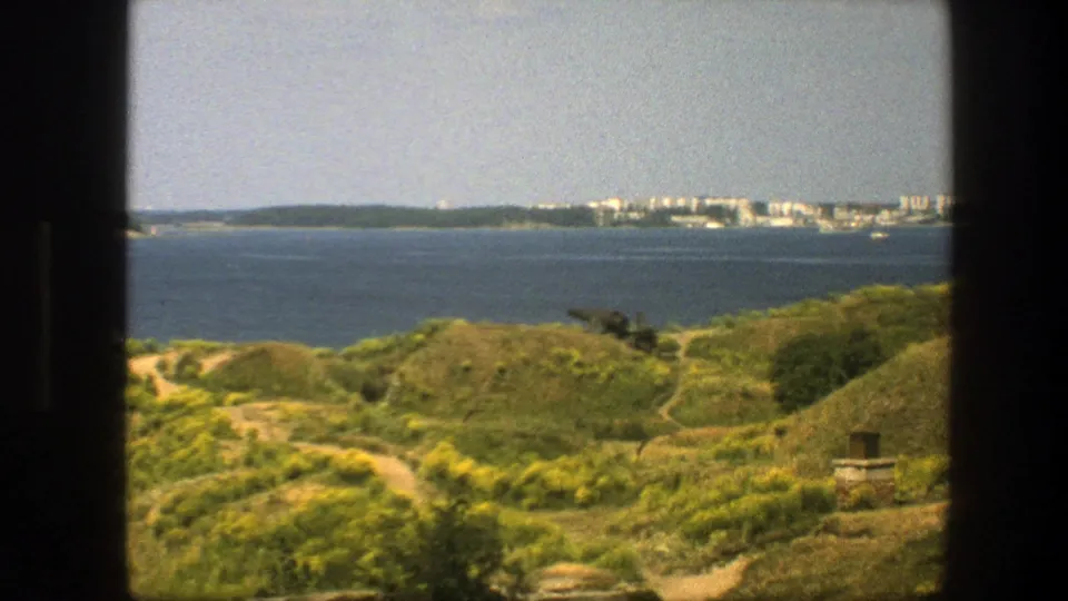 1975 Swedish 8mm frame of rolling coastal vegetation, intersecting paths, calm bay water, and a distant built-up shoreline.
