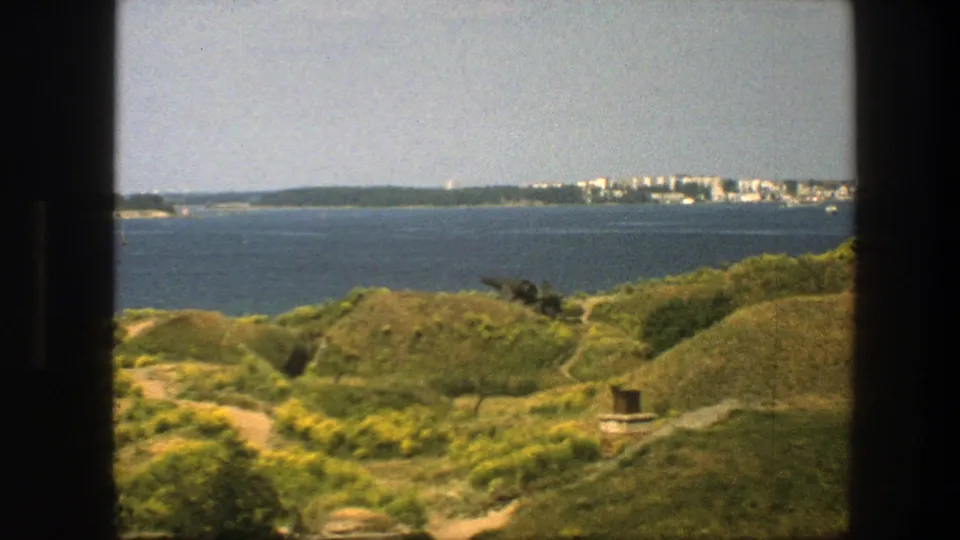 1975 Swedish coastal overlook with green hills, winding footpaths, a distant person on the slope, blue bay water, and far shoreline buildings.