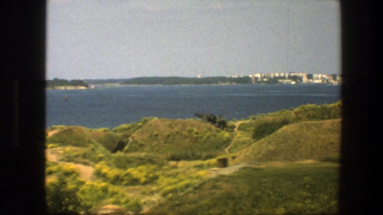 1975 8mm view in Sweden of green hills, wildflowers, blue sea, distant shoreline buildings, and a dark frame edge.