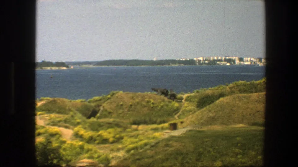 1975 8mm view in Sweden of green hills, wildflowers, blue sea, distant shoreline buildings, and a dark frame edge.