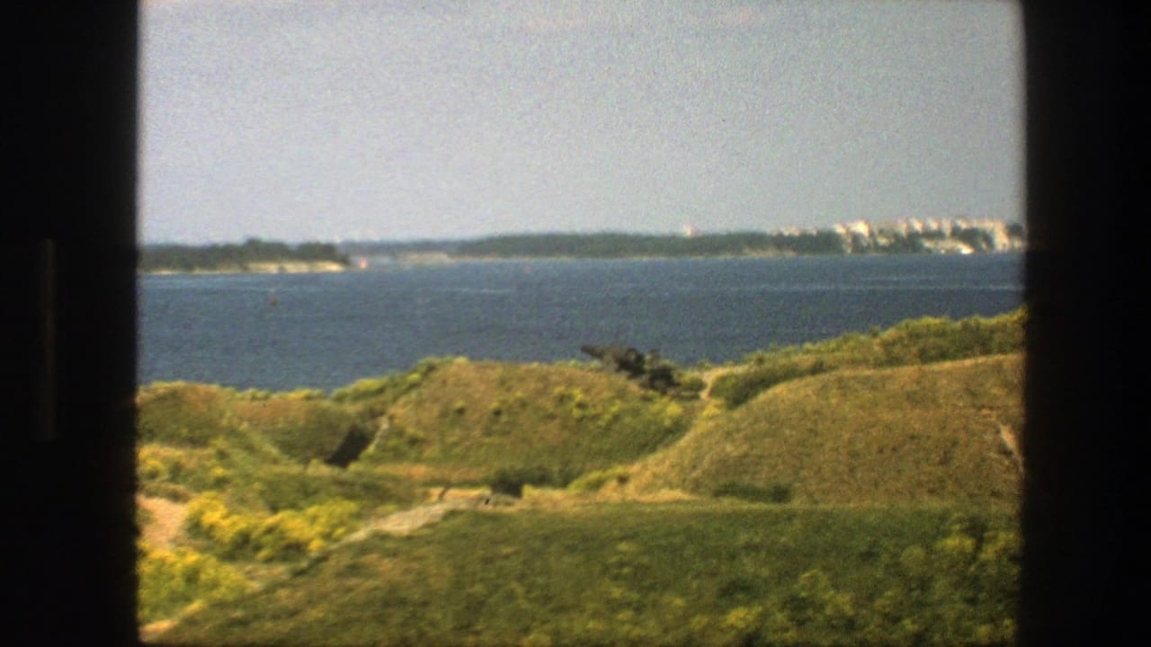 Sea view from grassy hills with distant shoreline buildings and dark vertical frame borders.