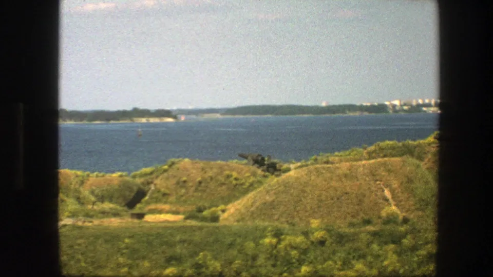 1975 8mm Swedish coastal scene with rolling grass, sea, distant shore, and what appears to be a dark cannon on the hill.