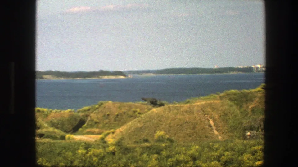 Swedish 1975 home-movie frame showing grassy coastal mounds, calm sea, distant buildings, and a possible cannon or artillery piece.