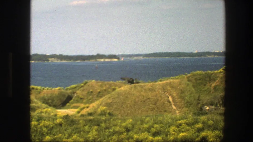 1975 Sweden home-movie view across blue coastal water and sunlit grassy hills from behind a dark window opening.