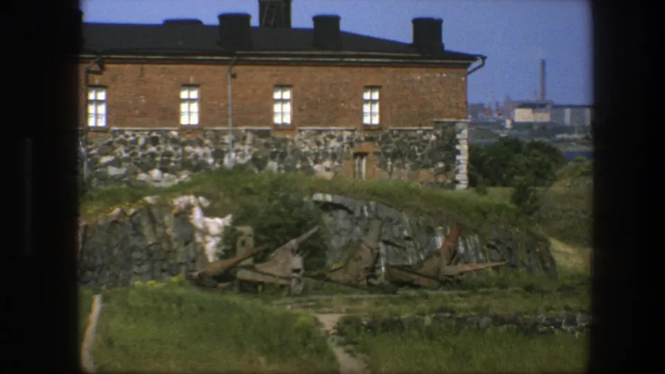 1975 Sweden home-movie frame showing a brick-and-stone building above grassy rocks and rusted artillery pieces.