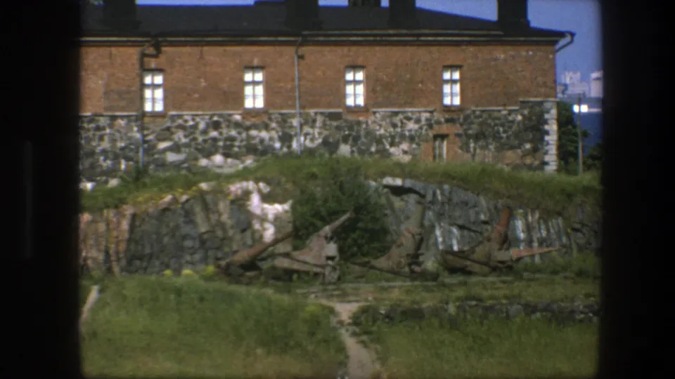 1975 Sweden 8mm view of a brick fortress structure on a stone base, seen across grass, rock, and weathered artillery remains.