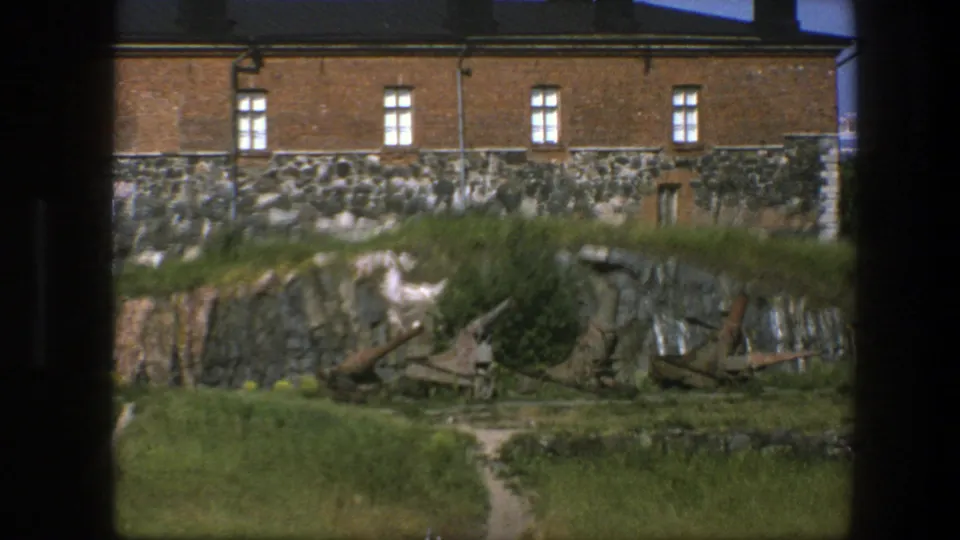 1975 Sweden 8mm frame of a coastal fortress building, rough stone base, grassy slope, narrow path, rock face, and rusted artillery pieces.