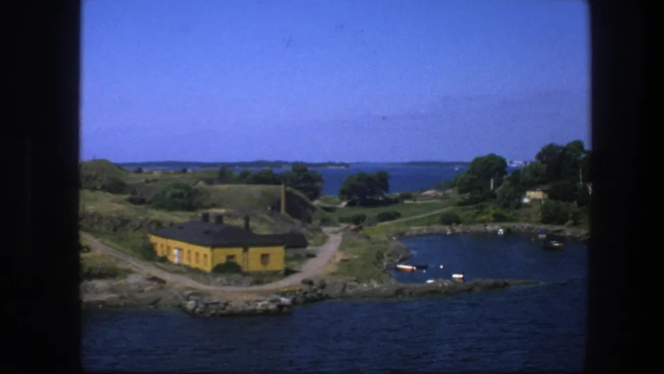 1975 Sweden 8mm harbor vista showing a yellow shoreline house, curving paths, blue water, small boats, trees, and distant islands.