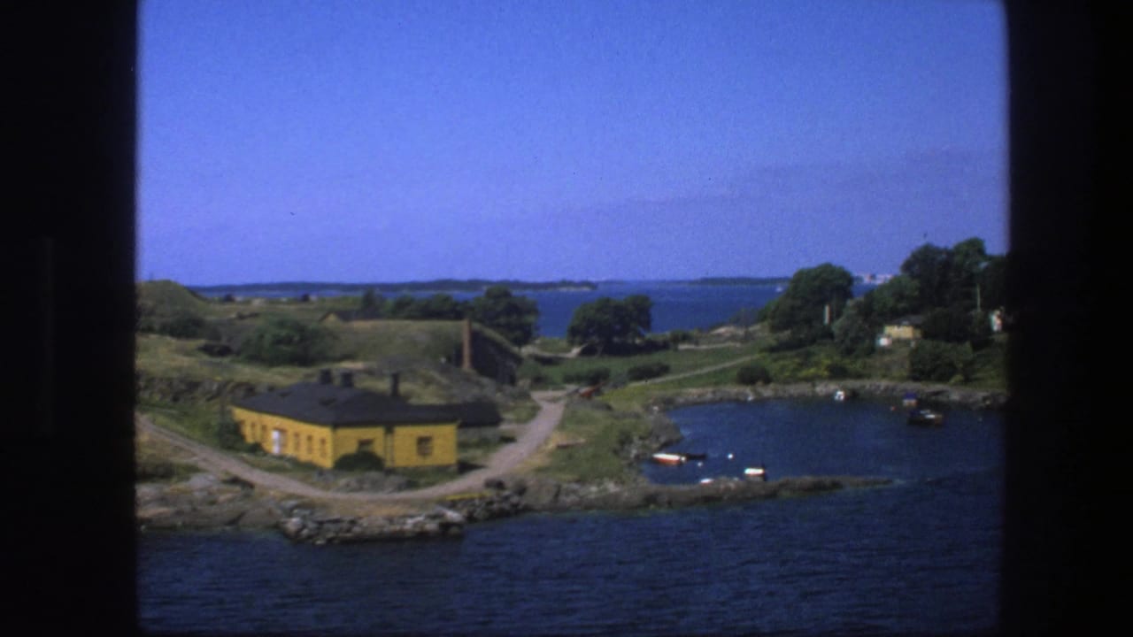 Coastal bay with yellow building, small boats, grassy shore, trees, and distant water.