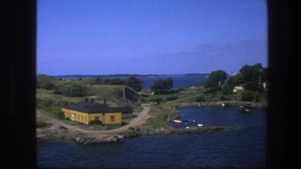 1975 Swedish 8mm view of a coastal bay with a yellow waterside building, small boats, green shore, and distant islands.