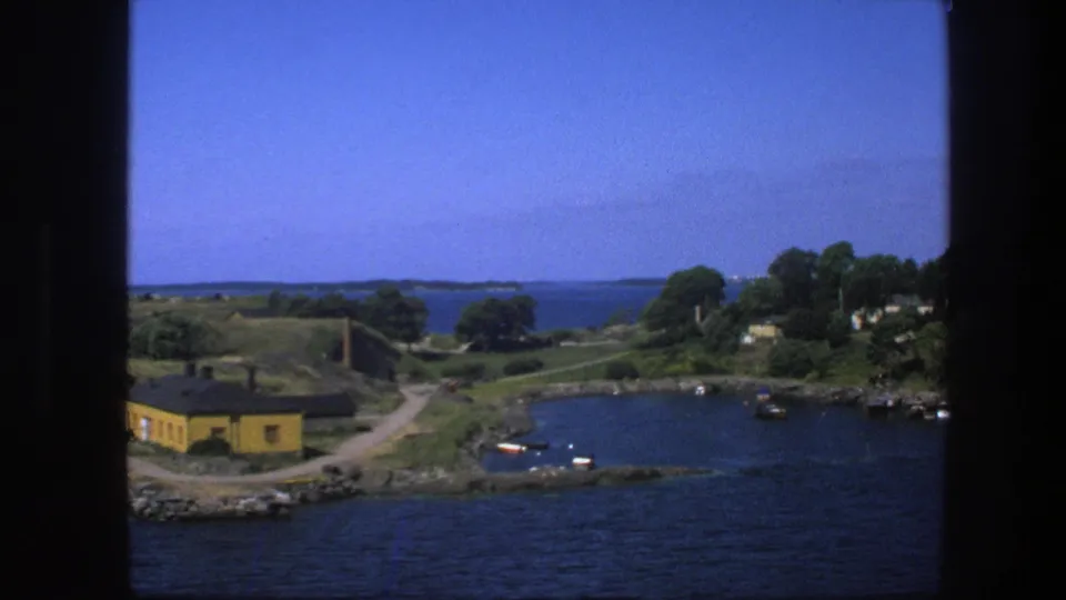 1975 Swedish 8mm shoreline scene with a yellow building, curved road, boats in a bay, tree-covered shore, and distant islands.