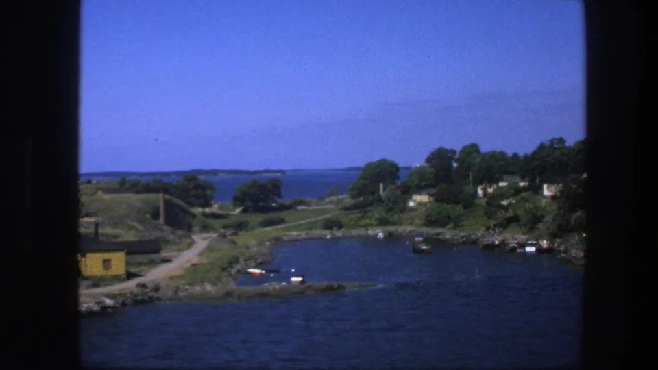 1975 Swedish coastal bay view with boats near the shore, a partly visible yellow building, green hillside, trees, and distant open water.