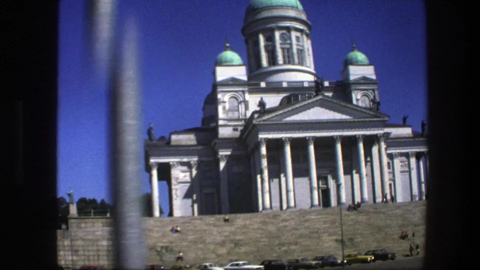 The cathedral-like building fills the frame, with white columns, green domes, people on the steps, and cars along the lower edge.