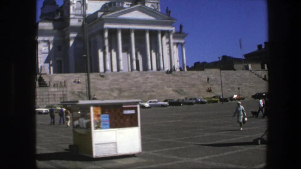 Sweden 1975 frame showing the cathedral entrance across the square, with a foreground kiosk, parked cars, and pedestrians walking through sunlight.